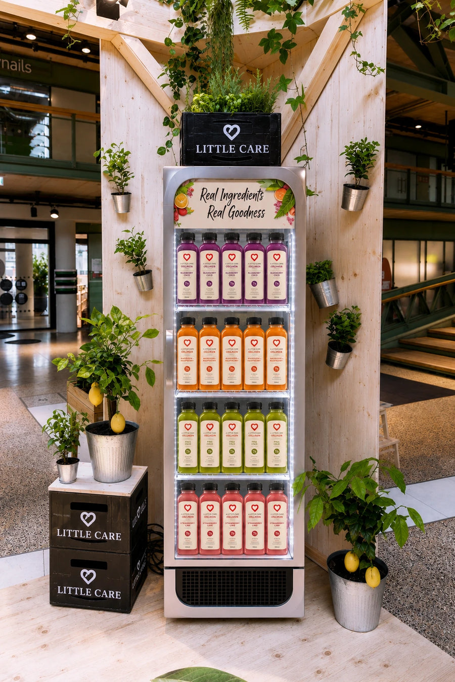 A tall Little Care beverage display refrigerator filled with colorful juice bottles, styled in a modern indoor space with wooden columns, potted plants, and lemon accents. The refrigerator has a black top sign and multiple shelves of pink, orange, yellow, and green drinks.